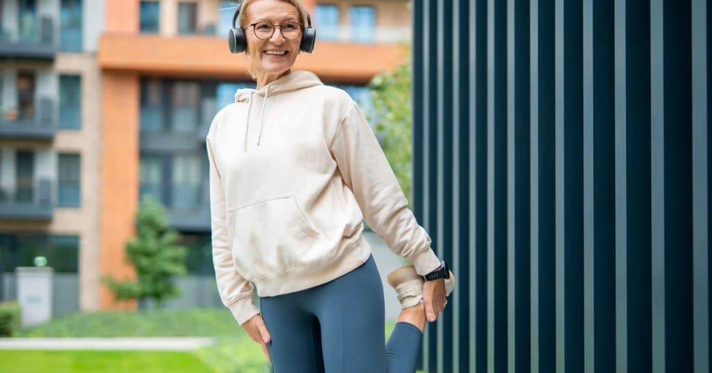 A woman smiles as she stretches her leg before or after exercise. She stands outdoors and wears headphones.