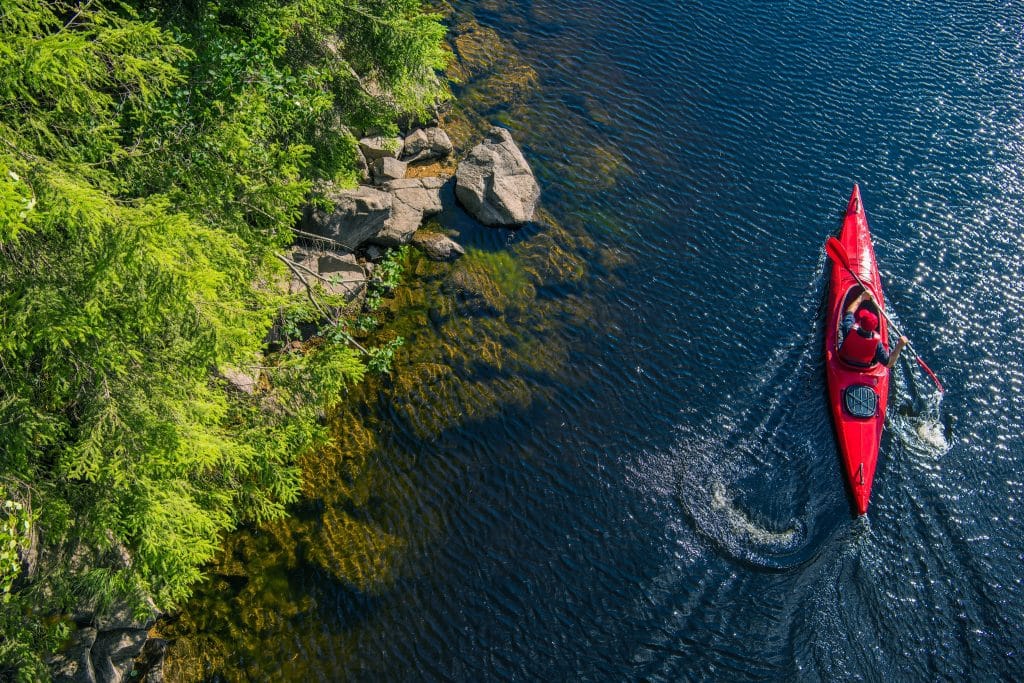 A high-angle view shows a person paddling a red kayak near a rocky, tree-covered shoreline on a sunny day.