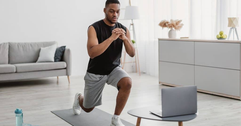 A person in athletic attire performing a lunge movement at home in front of an open small silver laptop.
