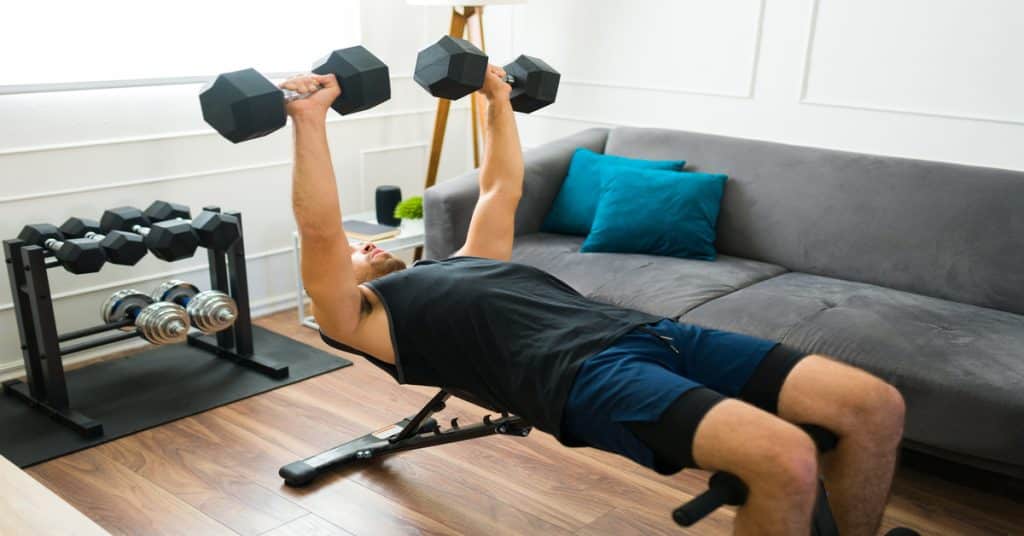 A man performs a dumbbell chest press on a weight bench in front of a gray couch with a set of weights behind him.