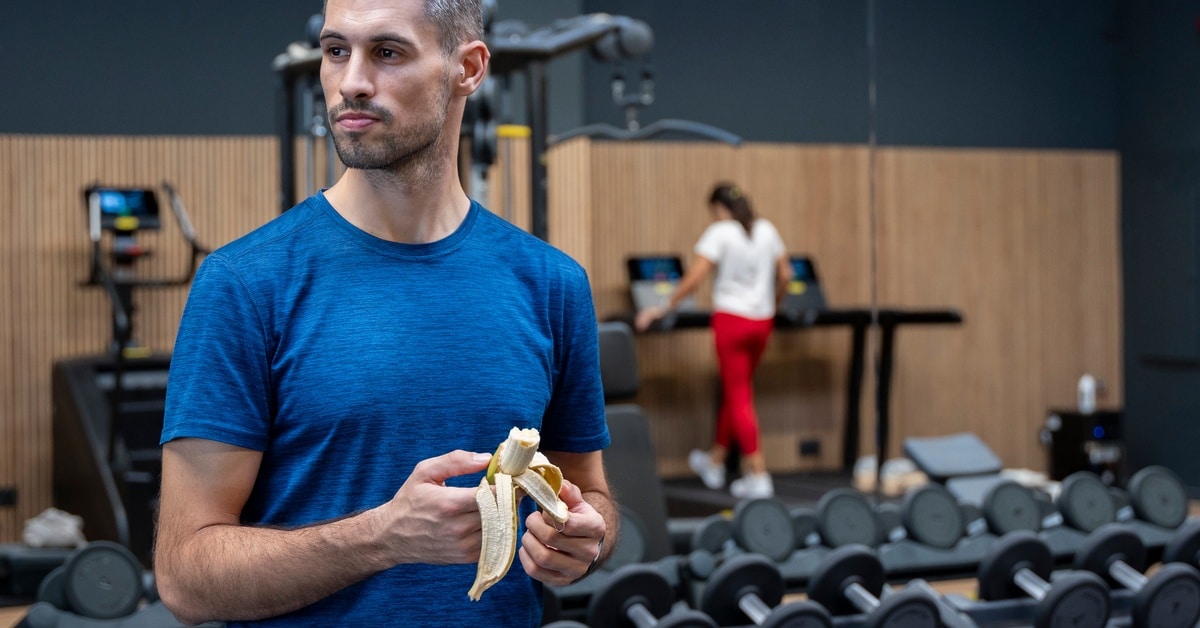 A man in a blue shirt holding a half-eaten banana while standing next to a full weight rack in a gym.