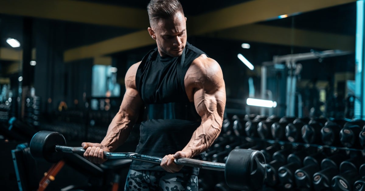 A young man in a sleeveless black top, curling a bar with weights in a modern gym with mirrors and weights behind him.