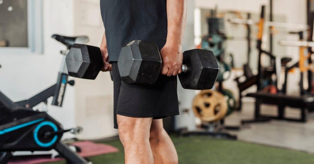 A man holds hex dumbbells at his sides in a farmer's carry position inside a home gym with equipment in the background.
