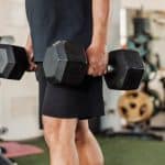 A man holds hex dumbbells at his sides in a farmer's carry position inside a home gym with equipment in the background.