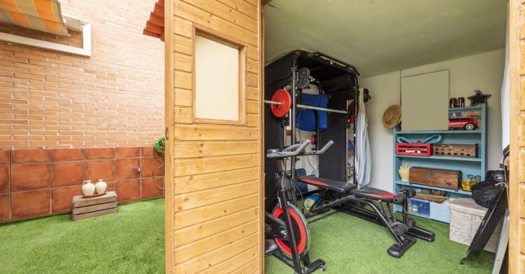 A backyard shed that has been converted into a small gym, featuring a squat rack, bench, and exercise bike.