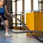 A senior woman in sportswear exercising with battle ropes inside a gym while bending forward during a workout.