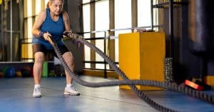 A senior woman in sportswear exercising with battle ropes inside a gym while bending forward during a workout.